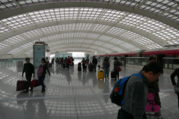 Menschen mit Gep├Ąck durch den Flughafen gehen, einige tragen Taschen und Koffer, mit einem Zug auf der rechten Seite und einer Glaswand im Hintergrund.