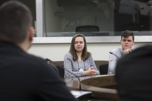 Eine Gruppe von Menschen sitzt um einen Tisch mit Papieren und einem Mikrofon, in eine Diskussion vertieft vor einem Glasfenster.