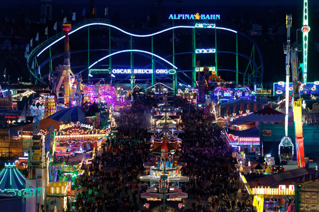 Eine große Menschenmenge steht um einen nächtlichen Karneval herum, der von bunten Lichtern beleuchtet wird und von Fahrgeschäften, Pfählen, Fahnen und Namensschildern umgeben ist, mit einem dunklen Hintergrund, der eine festliche Atmosphäre schafft.