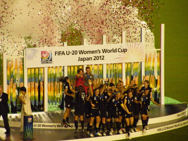 Eine Gruppe von Frauenfußballspielerinnen in schwarzen Uniformen feiert auf einer Bühne vor einer Menge, mit einer Tafel und dekorativen Gegenständen im Hintergrund.