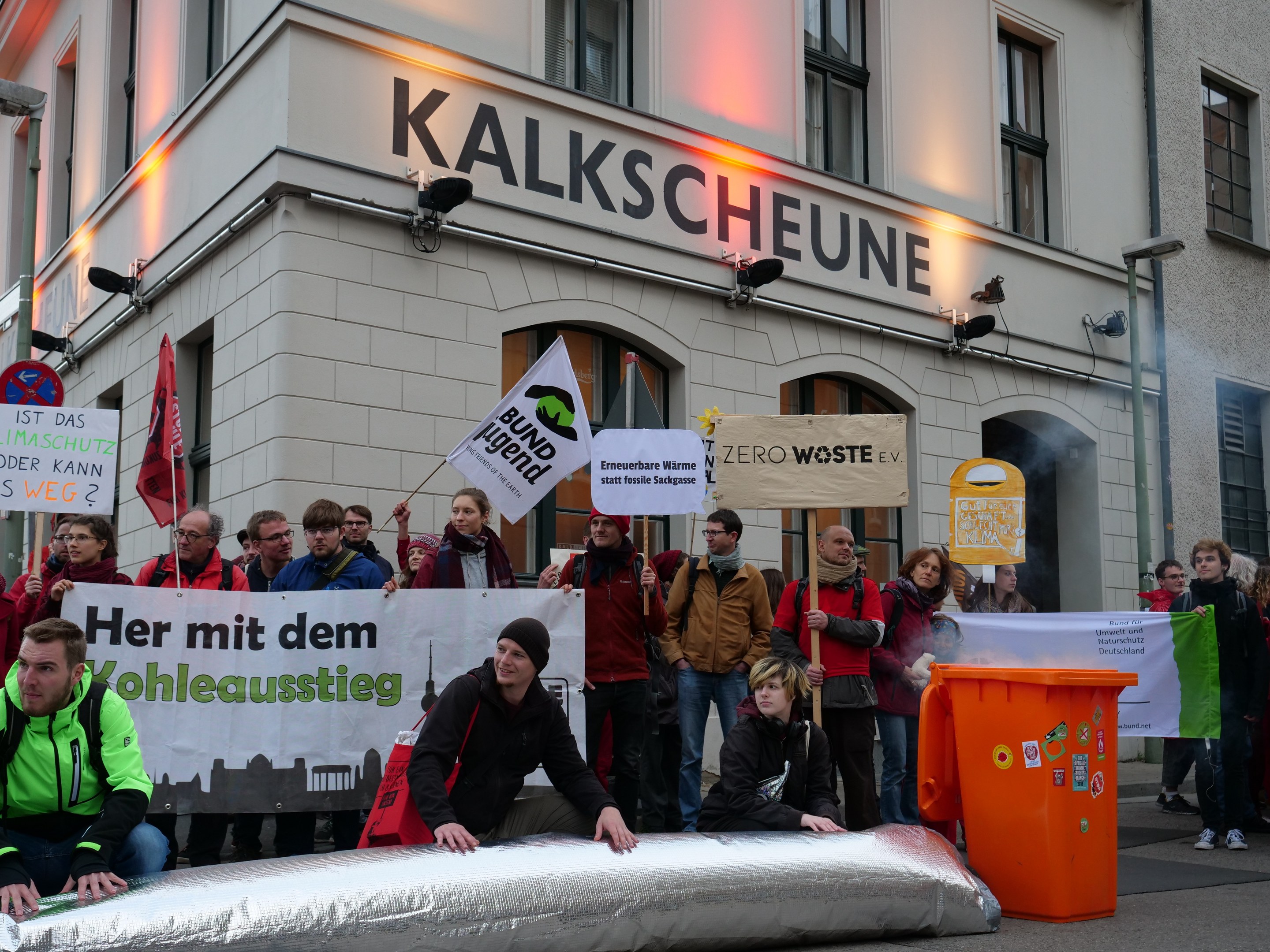 Gruppe von Menschen mit Schildern und Transparenten vor einem Gebäude während einer Demonstration in Deutschland, mit zwei Personen auf einem Objekt im Vordergrund und einem Müllcontainer auf der rechten Seite.
