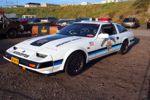 Ein Polizeiwagen auf einem Parkplatz neben anderen Fahrzeugen, Gras, Pfosten, Drähte, Gebäude und einen klaren blauen Himmel.