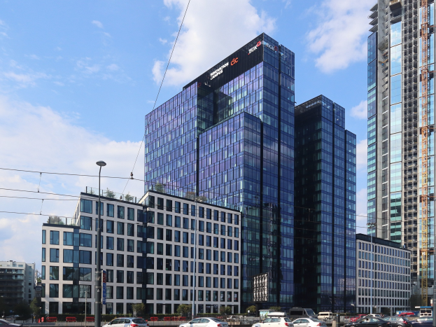 Großes modernes Gebäude mit vielen Fenstern, als Deutsche Bank Hauptquartier in Frankfurt, Deutschland identifiziert, umgeben von städtischer Infrastruktur einschließlich Straßenlaternen, Fahrzeugen, Bäumen und einem bewölkten Himmel.