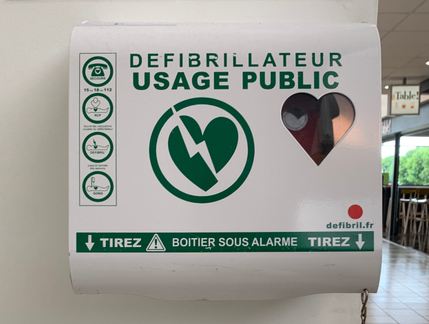 Ein Defibrillator-Gebrauchs-Schild an einer Gebäudewand, mit Stühlen, Tischen, Brettern, Lampen, Bäumen im Hintergrund und dem Boden unten.