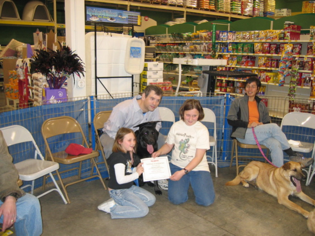 Eine Gruppe von Menschen sitzt auf Stühlen mit zwei Hunden, während zwei Kinder auf dem Boden knien und gemeinsam ein Zertifikat halten, mit Snacks, Lebensmitteln und Haushaltsgegenständen im Hintergrund.