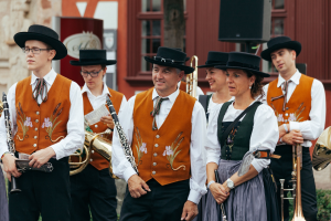 Eine Gruppe von Menschen in traditioneller bayrischer Tracht mit Musikinstrumenten, im Hintergrund ein Gebäude, Lautsprecher und Pflanzen.