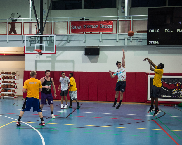 Eine Gruppe von Männern, die Basketball in einer Turnhalle spielen, mit einem Korb und Netz in der Mitte und einer Anzeigetafel auf der rechten Seite des Bildes.