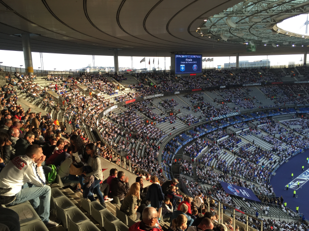 Eine große Menge von Menschen sitzt im Allianz Stadion in München, Deutschland, und schaut ein Fussballspiel, mit einer Bühne auf der rechten Seite, Fahnen, Mästen und einem Bildschirm im Hintergrund und der Himmel ist oben im Bild zu sehen.