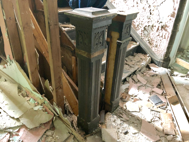 Ein Mann steht in einem renovierten Raum, umgeben von Werkzeugen und Materialien, mit einer Säule in der Mitte und Schutt auf dem Boden.