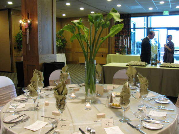 Ein Tisch mit einer weißen Decke, Tellern, Besteck, einem Glas und Baumzweigen, umgeben von Stühlen, mit einem grünen Tisch dahinter, einem Mann in einem schwarzen Blazer, einem stehenden Mädchen, einer Tür mit der Aufschrift "Ausgang" und zwei Männern hinter der Tür.