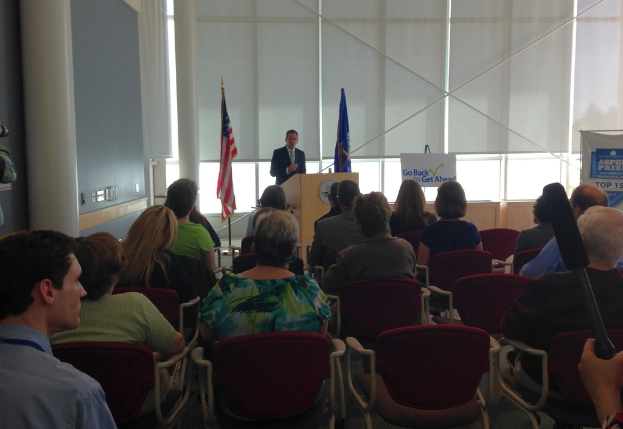 Ein Mann steht an einem Podium und spricht zu einem Publikum, das auf Stühlen sitzt, mit zwei Fahnen neben ihm und einer Wand, Säulen und Fenstern im Hintergrund.