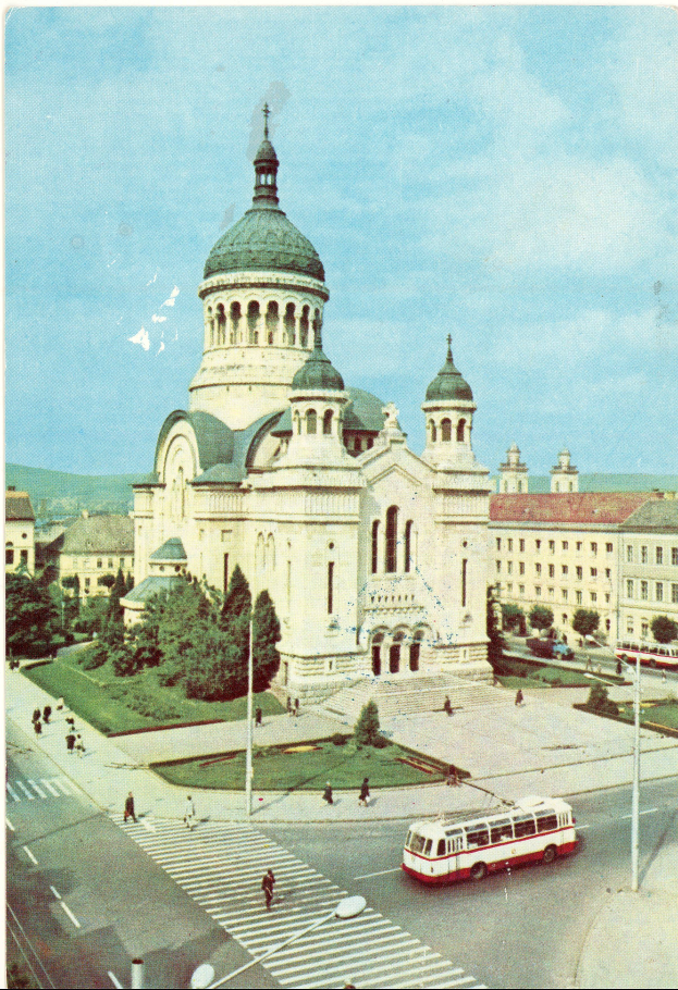 Ein altes foto einer stadtstraße mit einem bus, der daran vorbeifährt, umgeben von gebäuden, bäumen, masten, menschen, gras und einem klaren blauen himmel, mit text unten.