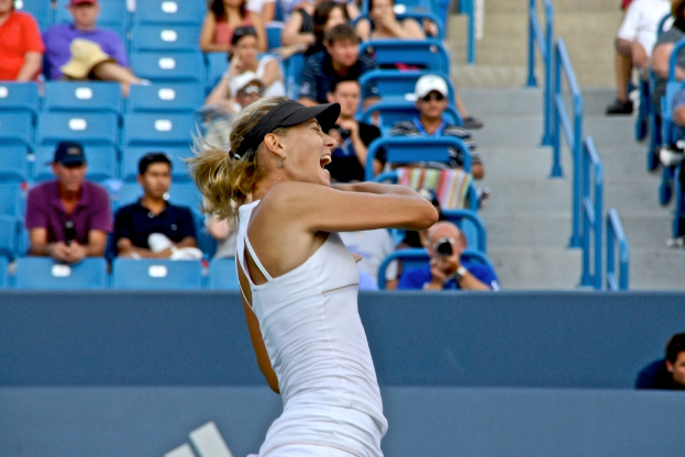 Eine Frau in weißem Oberteil und schwarzer Mütze steht vor einem blauen Stadionplatz mit sitzenden Zuschauern.