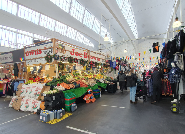 Ein belebter Indoor-Markt mit Menschen, die zwischen Obst- und Gemüse- sowie Kleiderständen gehen, unter Deckenlampen und in der Nähe großer Glasfenster.