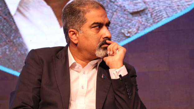 Ein Mann in Anzug und Krawatte sitzt auf einer Bühne und schaut nachdenklich auf einen Bildschirm.