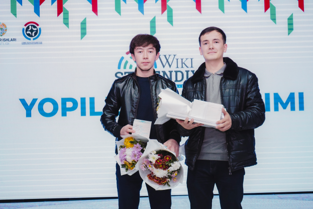 Zwei Männer in schwarzen Jacken mit Blumensträußen stehen auf einer Bühne mit einem "Yopil International Film Festival"-Banner im Hintergrund.