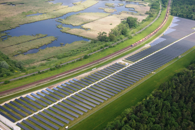 Luftaufnahme eines Solarparks mit Solarpanelen in einem Feld, umgeben von Bäumen, Gras, Pflanzen und Wasser, mit einem Zug auf einem nahen Bahngeleis.