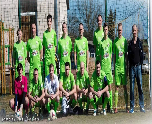 Gruppe von Männern in grünen T-Shirts auf einem Fußballfeld mit einem Ball, einige in Hockstellung, in der Nähe eines Torpfostens, mit Bäumen, Gebäuden und Fahrzeugen im Hintergrund.