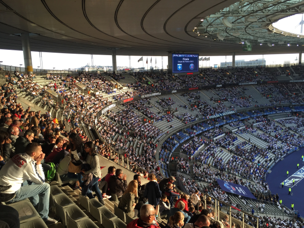 Große Menschenmenge im Allianz Arena Stadion in München, Deutschland, bei einem Fußballspiel mit einer Bühne, Flaggen, Stangen und einem Bildschirm im Hintergrund.