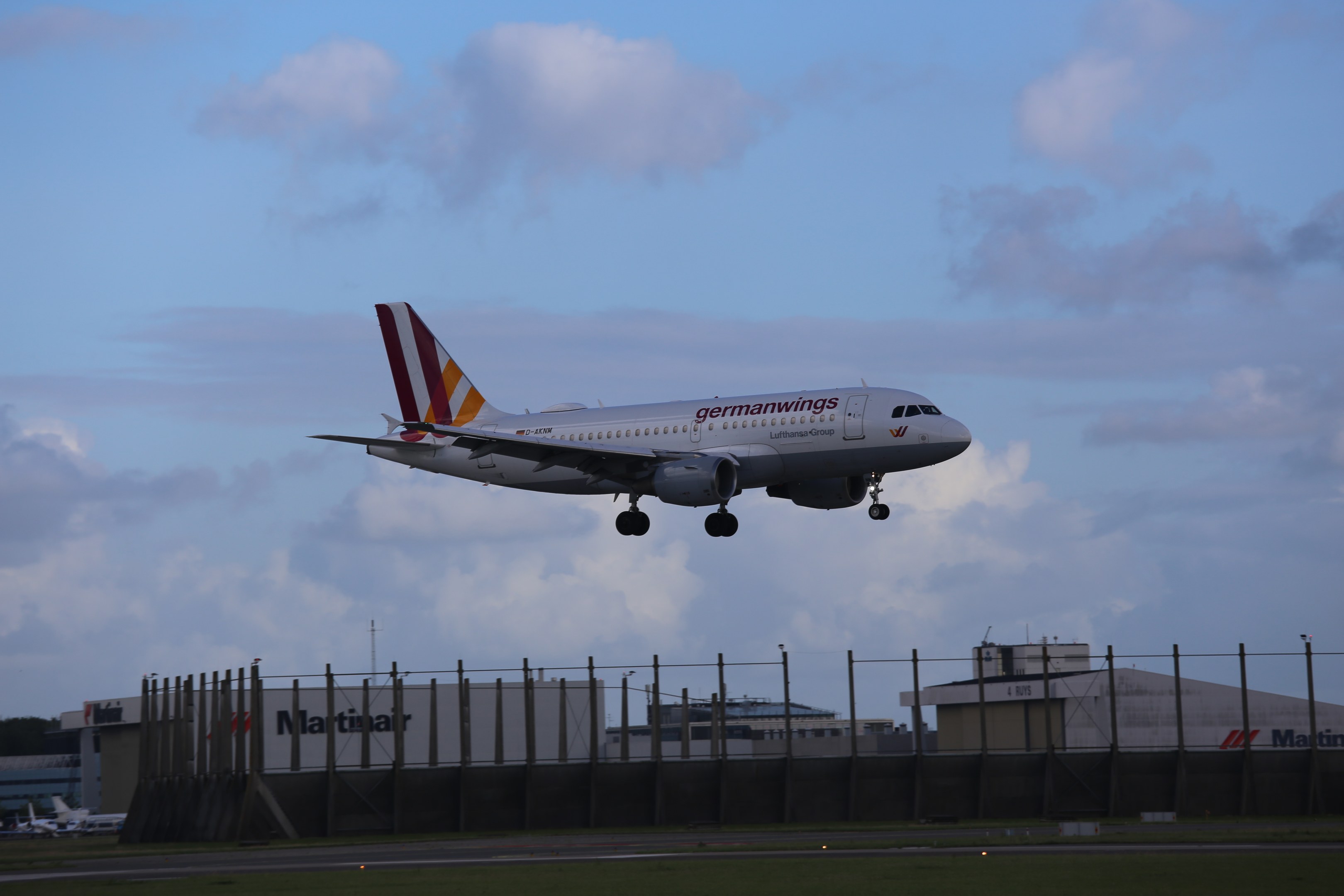 Germanwings Airbus A320-200 beim Abheben vom Frankfurter Airport mit Gras darunter, einem Zaun im Vordergrund, Gebäuden und Bäumen im Hintergrund und einer bewölkten Himmel über.