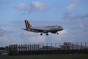 Germanwings Airbus A320-200 beim Abheben vom Frankfurter Airport mit Gras darunter, einem Zaun im Vordergrund, Gebäuden und Bäumen im Hintergrund und einer bewölkten Himmel über.
