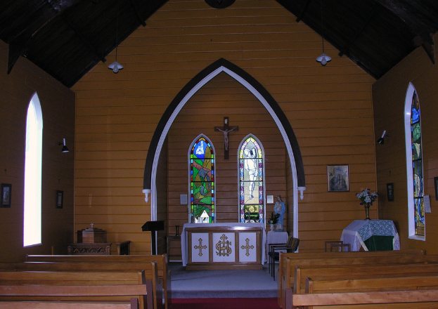 Das Bild zeigt das Innere einer Kirche mit Bänken auf beiden Seiten, einem Kreuz in der Mitte, hängenden Lampen oben, einem Tisch mit einer Blumenvase und einem gerahmten Bild auf der rechten Seite und einem zentralen Idol, das von einem Tisch mit einem Tuch flankiert wird.