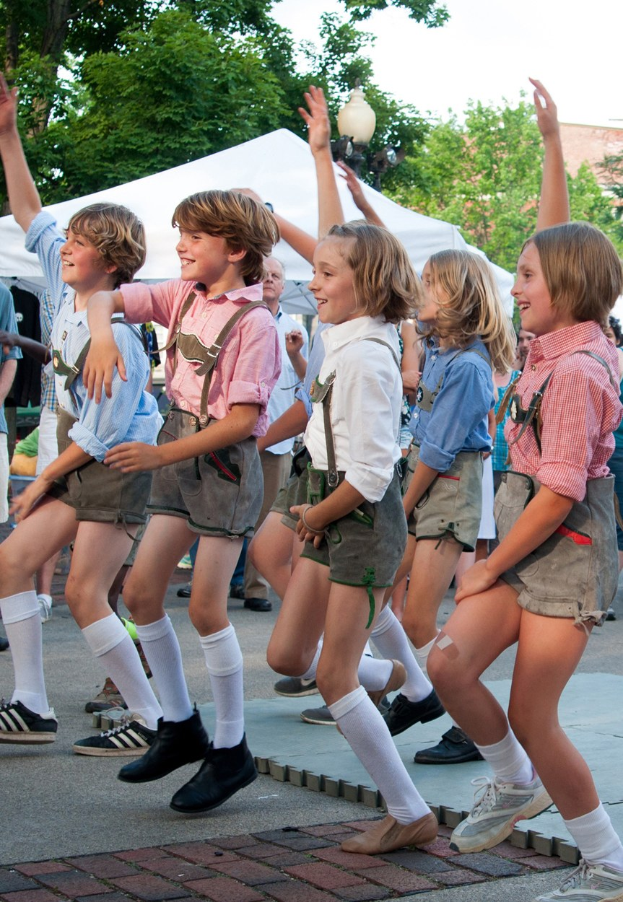 Kinder in traditioneller bayrischer Kleidung tanzen auf einer Straße, umgeben von einer Menge, Zelten, Laternenmasten, Bäumen, Gebäuden und einem klaren blauen Himmel.