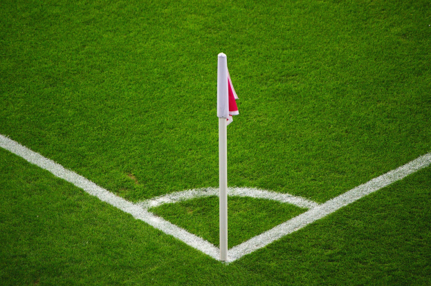 Fussballfeld mit gruener Wiese, weissen Begrenzungslinien und einem weissen Pfahl mit einer rot-weissen Flagge, die den Elfmeterpunkt markiert.