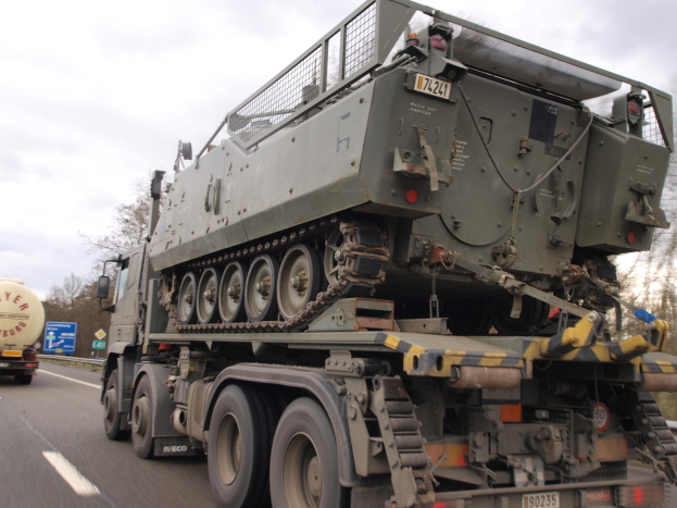 Militärlastwagen transportiert einen Panzer auf einer Autobahn mit Bäumen, Holzplanken und einem klaren blauen Himmel im Hintergrund.
