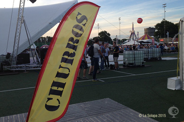 Ein buntes Volksfest-Szenario mit Zelten, einem zentralen Mast, Menschen, Bäumen, Strommasten, Straßenbahnen auf der Straße und einem eisernen Stand auf der linken Seite unter einem klaren Himmel.