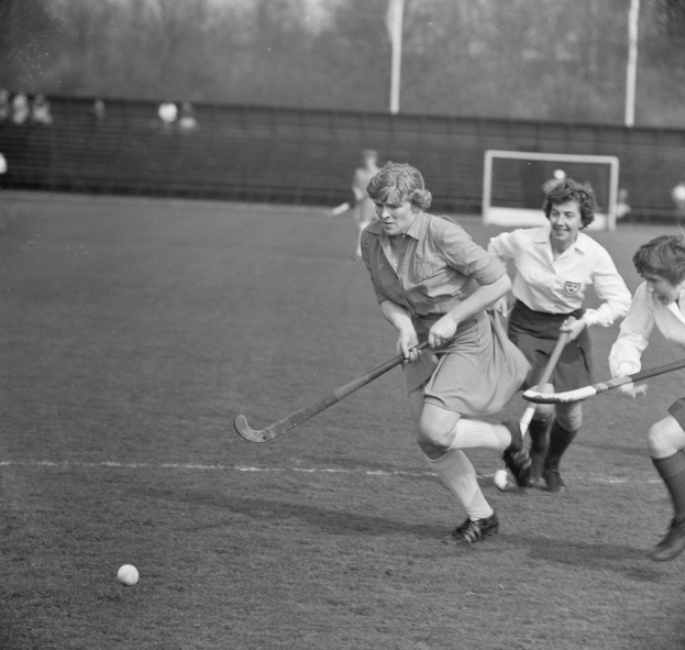 Eine Gruppe von Frauen, die Hockey auf dem Feld spielen, mit einem Ball in der Mitte der Aktion, einem Torpfosten, einer Wand, Pfählen, Bäumen und einigen Zuschauern im Hintergrund, in Schwarz-Weiß.