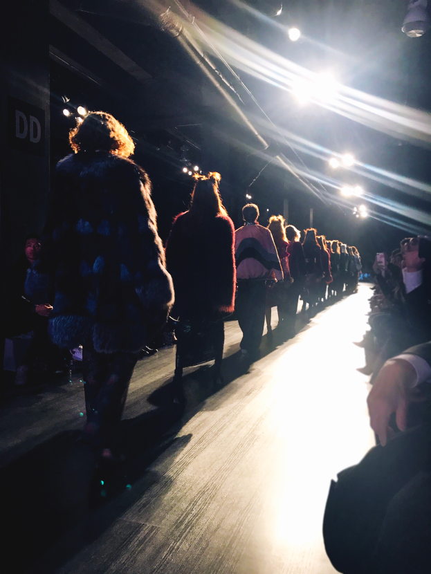 Eine Gruppe von Menschen läuft auf einem hell beleuchteten Laufsteg bei einer Modenschau, mit Zuschauern auf beiden Seiten.