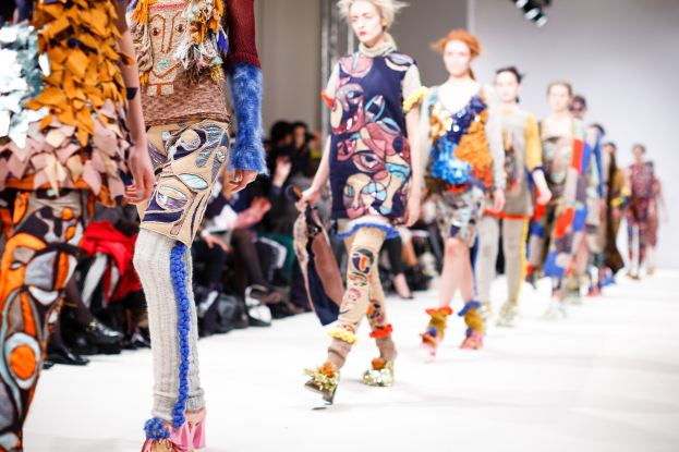 Eine Gruppe von Frauen, die auf dem Laufsteg der London Fashion Week gehen, mit sitzenden Zuschauern und Bühnenbeleuchtung im Hintergrund.