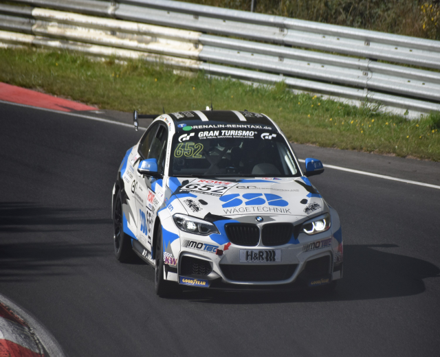 Ein BMW M4 DTM-Rennwagen auf einer Rennstrecke, umgeben von Gras, einem Zaun und Pflanzen, mit sichtbarem Text, Zahlen und einem Rad.