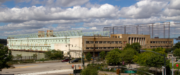 Eine breite Straße mit Fahrzeugen, gesäumt von Bäumen und Straßenlaternen, führt zu einem großen architektonischen Gebäude in der Nähe eines Gewässers.
