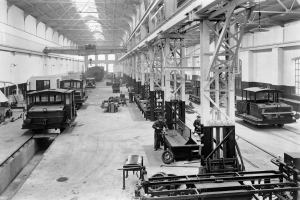 Ein Schwarz-Weiß-Foto einer Fabrikhalle mit mehreren Zügen auf dem Boden, Arbeitern, Metallstangen, Deckenlampen und Wandfenstern.