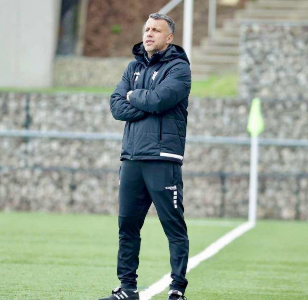 Ein Fußballtrainer auf einem Feld während einer Trainingseinheit, die Arme verschränkt, mit einer schwarzen Jacke und Hose, vor einem Zaun, Treppe und Wand.