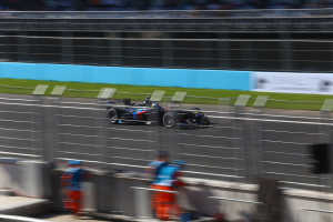 Ein Formelwagen fährt auf einer Rennstrecke mit einer unscharfen Zuschauermenge am Straßenrand, Gras am Boden und Tafeln mit Text im Hintergrund.