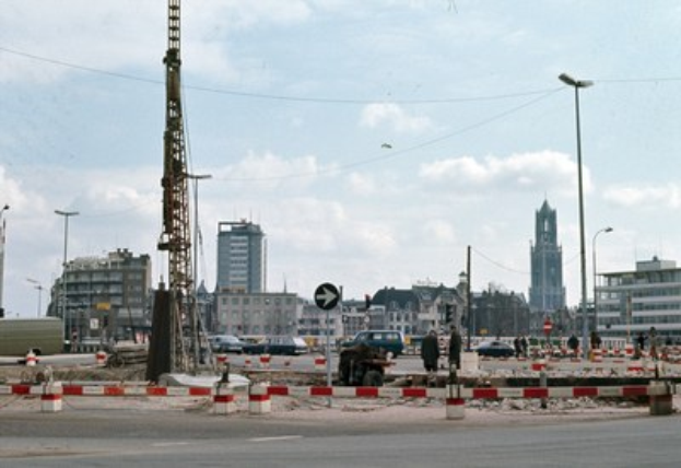 Baustelle mit einem zentralen Turm, umgeben von Gebäuden, Straßeninfrastruktur, Fahrzeugen und bewölktem Himmel.