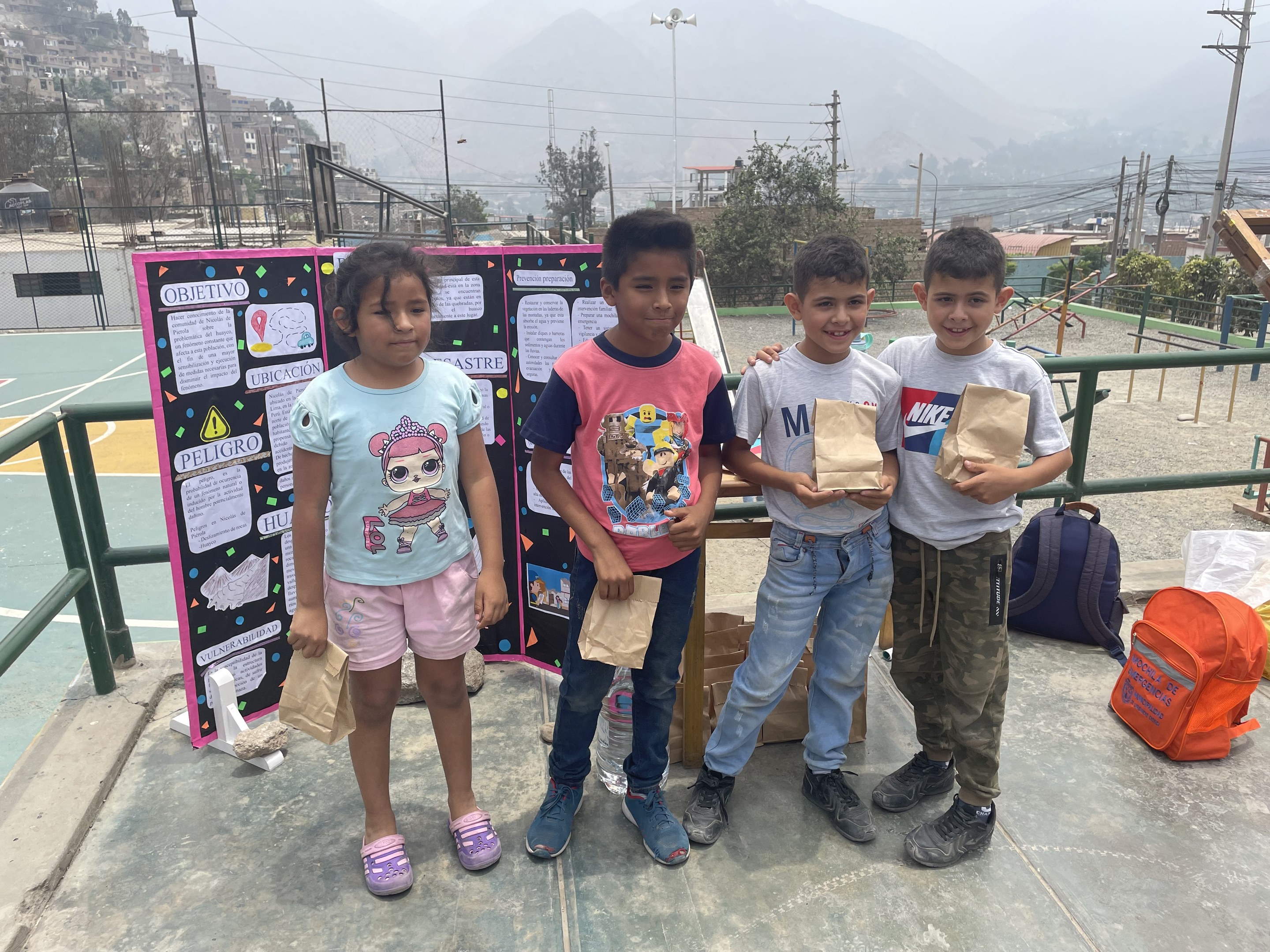 Gruppe von Kindern auf einem Basketballfeld mit Papierbeuteln in der Hand, mit Beuteln und Gegenständen rechts und einer Tafel mit Text links, vor einem Hintergrund aus Zaun, Pfosten, Straßenlaternen, Bäumen, Gebäuden, Hügeln und Himmel.