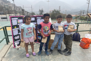 Gruppe von Kindern auf einem Basketballfeld mit Papierbeuteln in der Hand, mit Beuteln und Gegenständen rechts und einer Tafel mit Text links, vor einem Hintergrund aus Zaun, Pfosten, Straßenlaternen, Bäumen, Gebäuden, Hügeln und Himmel.