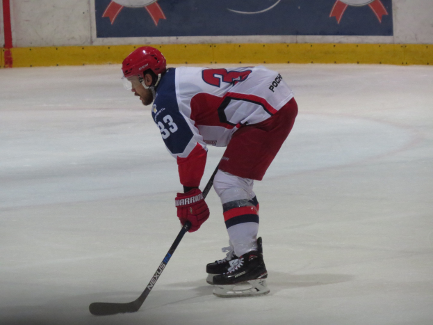 Ein Hockey-Spieler in voller Ausrüstung fährt auf dem Eis mit einem Stock, der einer Wand mit Banner im Hintergrund zugewandt ist.