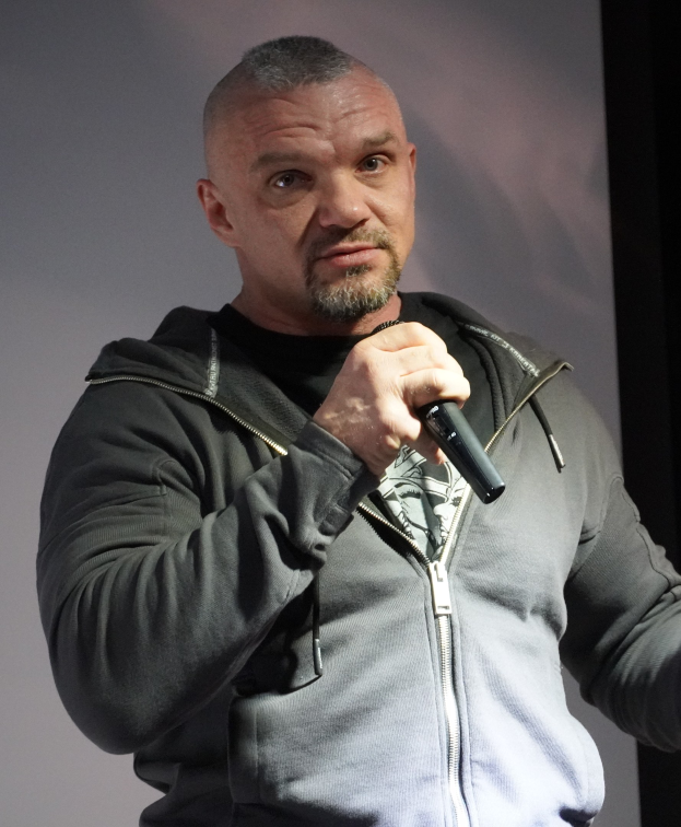 Ein Mann in einer schwarzen Kapuzenjacke und einem schwarzen Anzug mit weißem Hemd hält ein Mikrofon und steht selbstbewusst vor einer Wand, was darauf hindeutet, dass er ein Komiker ist.