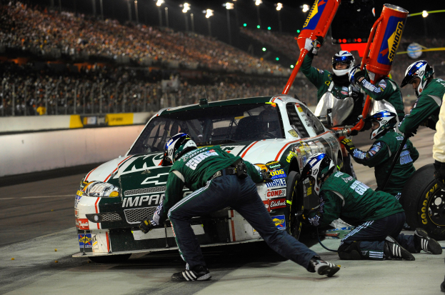 NASCAR-Auto wird von Crewmitgliedern in Helmen mit einem beleuchteten Stadion im Hintergrund gewartet.