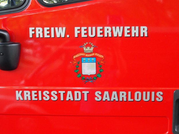 Ein roter Feuerwehrauto mit den Worten "Freiwillige Feuerwehr Landkreis Saarlouis" auf der Seite, mit einer Tür mit Griff und Fenstern, die Bäume im Hintergrund zeigen.