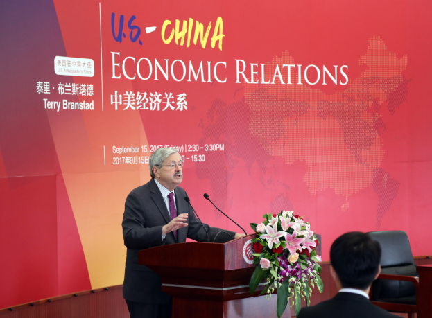 Ein Mann in einer schwarzen Jacke, weißem Hemd und lila Krawatte steht an einem Podium mit zwei Mikrofonen und einem Blumenstrauß und spricht vor einer roten Wand, auf der 'US-China Economic Relations' steht, mit einer Person rechts daneben sitzend.