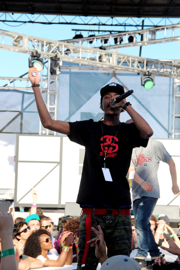 Eine Person in einem schwarzen T-Shirt hält ein Mikrofon auf einer Bühne mit Menschen drumherum, und eine weitere Person in einem grauen T-Shirt steht im Hintergrund unter einem blauen Himmel.