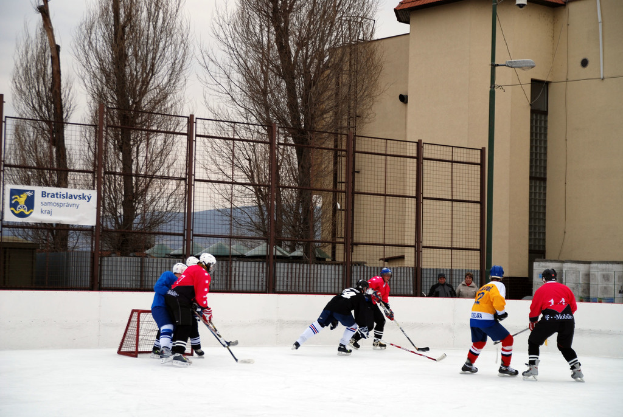 Menschen, die Eis Hockey auf einem Eisplatz mit Gebäuden, Bäumen, einer Straßenlaterne, einer Namensschilder und Zäunen im Hintergrund unter einem Himmel spielen.