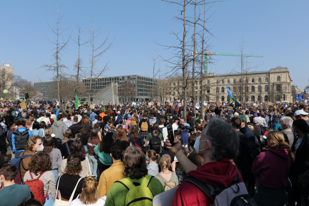 Eine große Menge Menschen mit Schildern und Taschen steht vor einem Gebäude mit Fenstern, Bäumen und einem klaren Himmel während einer Klimaprotest.