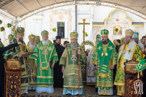 Eine Gruppe von Priestern steht vor einer Kirche während einer religiösen Zeremonie, wobei einer ein Buch und ein Mikrofon hält, während Blumen, ein Kreuz und ein Gebäude im Hintergrund zu sehen sind.